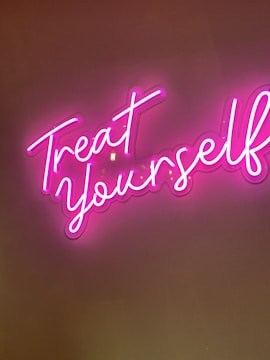 A neon sign with cursive handwriting displaying the words 'Treat Yourself' in bright pink against a dark background.