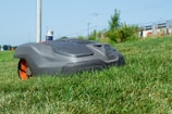 A robotic lawn mower is moving across a grassy lawn, with a clear blue sky in the background. The mower has a sleek, compact design and features prominent black and orange details.