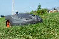 A robotic lawn mower is moving across a grassy lawn, with a clear blue sky in the background. The mower has a sleek, compact design and features prominent black and orange details.