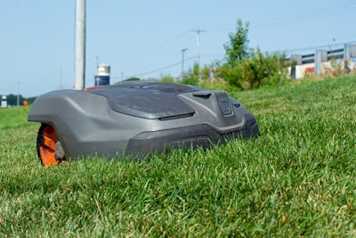 A robotic lawn mower is moving across a grassy lawn, with a clear blue sky in the background. The mower has a sleek, compact design and features prominent black and orange details.