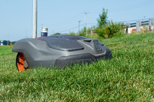 A robotic lawn mower is moving across a grassy lawn, with a clear blue sky in the background. The mower has a sleek, compact design and features prominent black and orange details.