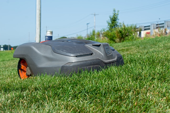 A robotic lawn mower is moving across a grassy lawn, with a clear blue sky in the background. The mower has a sleek, compact design and features prominent black and orange details.