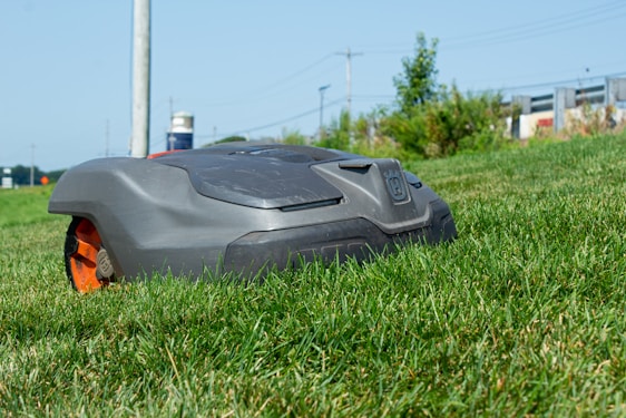 A robotic lawn mower is moving across a grassy lawn, with a clear blue sky in the background. The mower has a sleek, compact design and features prominent black and orange details.