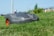 A robotic lawn mower is moving across a grassy lawn, with a clear blue sky in the background. The mower has a sleek, compact design and features prominent black and orange details.