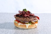 A gourmet burger featuring a thick beef patty topped with crispy bacon strips and a small serving of greens. The burger includes layers of fresh tomato slices, pickles, red onion rings, and creamy sauce, all sandwiched between a soft bun.