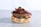 A gourmet burger featuring a thick beef patty topped with crispy bacon strips and a small serving of greens. The burger includes layers of fresh tomato slices, pickles, red onion rings, and creamy sauce, all sandwiched between a soft bun.
