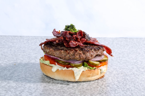 A gourmet burger featuring a thick beef patty topped with crispy bacon strips and a small serving of greens. The burger includes layers of fresh tomato slices, pickles, red onion rings, and creamy sauce, all sandwiched between a soft bun.