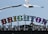 Brighton Palace Pier sign with colorful, illuminated letters against a clear blue sky. Two seagulls are flying above, and another is perched on top of the sign.