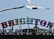 Brighton Palace Pier sign with colorful, illuminated letters against a clear blue sky. Two seagulls are flying above, and another is perched on top of the sign.
