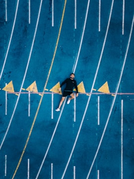 Aerial view of a person lying on a blue running track with marked lanes. Yellow triangular markers and a yellow line intersect the track. The person is dressed in black with white shoes.