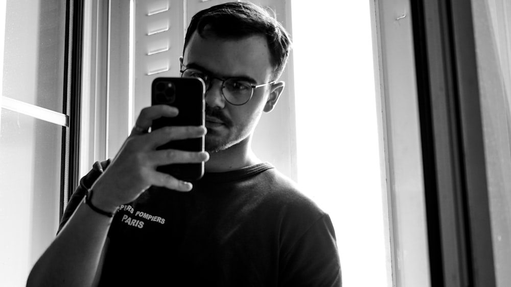man in black crew neck shirt wearing black framed eyeglasses holding black smartphone