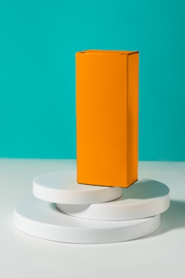 A tall, rectangular orange box stands upright on top of three stacked white circular platforms, set against a vibrant turquoise background.