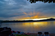 A serene lakeside view during sunset, with the sun&rsquo;s rays casting a golden reflection on the water. Clouds partially obscure the sky, adding depth and drama to the scene. Boats and loungers are seen along the water&rsquo;s edge, while trees and hills form a dark silhouette in the background.