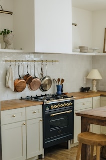 A kitchen area featuring a dark-colored stove with brass accents, surrounded by light cream cabinetry and wooden countertops. Copper and silver pots hang from a brass rail above the stove. A wooden table and stool are in the foreground, and there are decorative items like a blue vase and utensils on the counter.