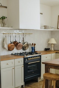 A kitchen area featuring a dark-colored stove with brass accents, surrounded by light cream cabinetry and wooden countertops. Copper and silver pots hang from a brass rail above the stove. A wooden table and stool are in the foreground, and there are decorative items like a blue vase and utensils on the counter.