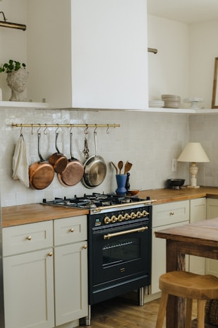 A kitchen area featuring a dark-colored stove with brass accents, surrounded by light cream cabinetry and wooden countertops. Copper and silver pots hang from a brass rail above the stove. A wooden table and stool are in the foreground, and there are decorative items like a blue vase and utensils on the counter.