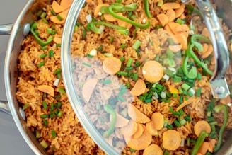 A close-up view of a skillet filled with cooked rice mixed with sliced carrots, diced green onions, and thin strips of green bell peppers. The skillet is covered with a glass lid, partially fogged, giving a glimpse of the vibrant colors of the ingredients.