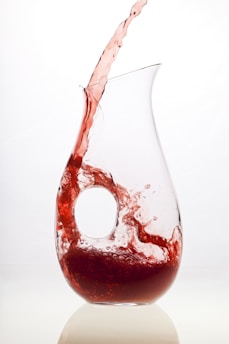 A clear glass pitcher with a unique circular handle is shown with red liquid being poured into it. The liquid creates a dynamic splash effect, partially filling the lower portion of the pitcher. The background is a plain white, enhancing the contrast with the red liquid.