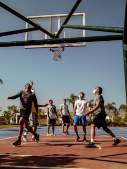 Several individuals are playing basketball outdoors on a sunny day. They are wearing casual sportswear and face masks, suggesting a setting influenced by health precautions. The basketball hoop is prominently featured against a clear blue sky. The surrounding area includes palm trees and greenery.
