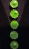 Five green circular objects arranged vertically on a dark surface. The objects have a spiked outer edge, resembling cupcake liners. Light casts subtle shadows on the surface, highlighting the texture.
