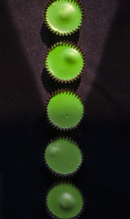 Five green circular objects arranged vertically on a dark surface. The objects have a spiked outer edge, resembling cupcake liners. Light casts subtle shadows on the surface, highlighting the texture.