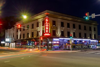 A brightly lit corner building at night featuring a bar with a neon sign. Several cars pass by, leaving light trails. The streets are illuminated by traffic lights, with a mix of red, green, and amber signals. The building has multiple floors with windows and contains signage indicating an open patio and bar.