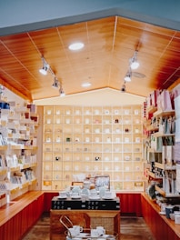 A well-organized store interior featuring a variety of neatly arranged mugs on shelves that cover the back wall. The shelves are illuminated, highlighting the colorful and diverse mug designs. On the side walls, additional shelves display stationery items and gift bags. The center of the room has a wooden counter displaying tableware and kitchen accessories.