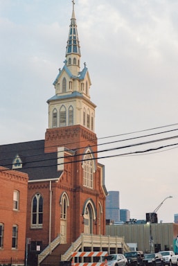 A historic brick church with a tall steeple rising against a cloudy sky. The building features arched windows and steps leading up to the entrance. Surrounding the church are other urban structures and a few parked cars, with power lines crossing the scene.