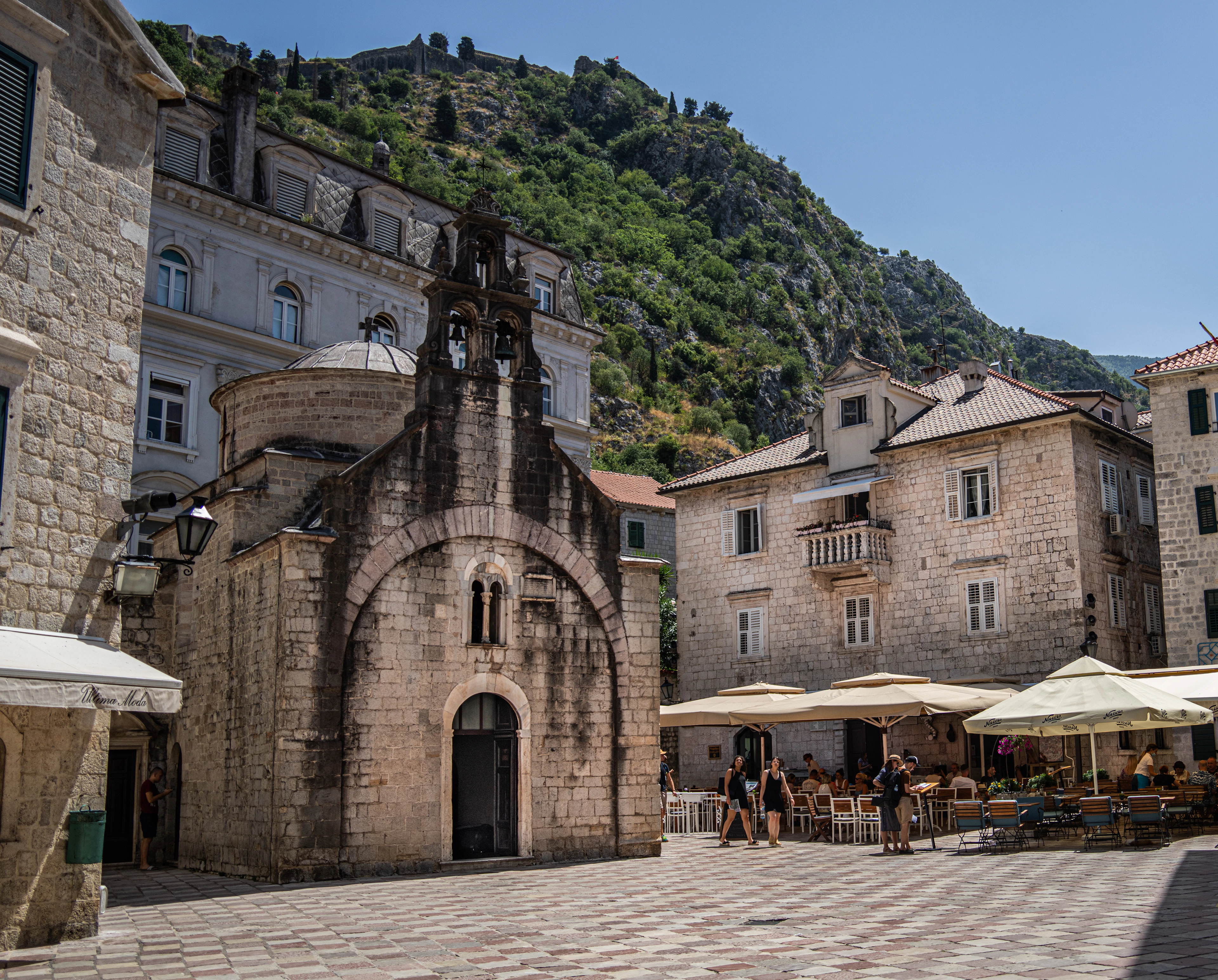 A small Romanesque stone church and cafe umbrellas on a paved square in Kotor's old town, under the green cliff