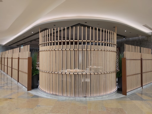 A modern, open-space interior featuring a circular structure made of parallel wooden slats, creating a semi-enclosed area. The design exudes a minimalist aesthetic with a focus on natural materials. The surrounding area includes tall, green plants placed in large pots, adding a touch of nature to the space. The ceiling is elegantly curved with recessed lighting, while the floor is made of polished stone tiles.