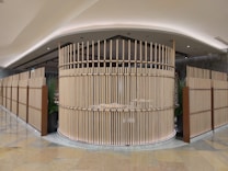 A modern, open-space interior featuring a circular structure made of parallel wooden slats, creating a semi-enclosed area. The design exudes a minimalist aesthetic with a focus on natural materials. The surrounding area includes tall, green plants placed in large pots, adding a touch of nature to the space. The ceiling is elegantly curved with recessed lighting, while the floor is made of polished stone tiles.