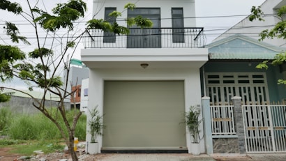 The image depicts a modern two-story house with a white facade and a balcony on the upper level. There is a large garage door in the center of the lower level. A small tree with green leaves is visible on the left side, and some grass grows near it. The house has a clean and minimalistic design.