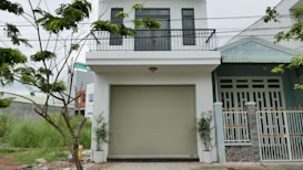 The image depicts a modern two-story house with a white facade and a balcony on the upper level. There is a large garage door in the center of the lower level. A small tree with green leaves is visible on the left side, and some grass grows near it. The house has a clean and minimalistic design.
