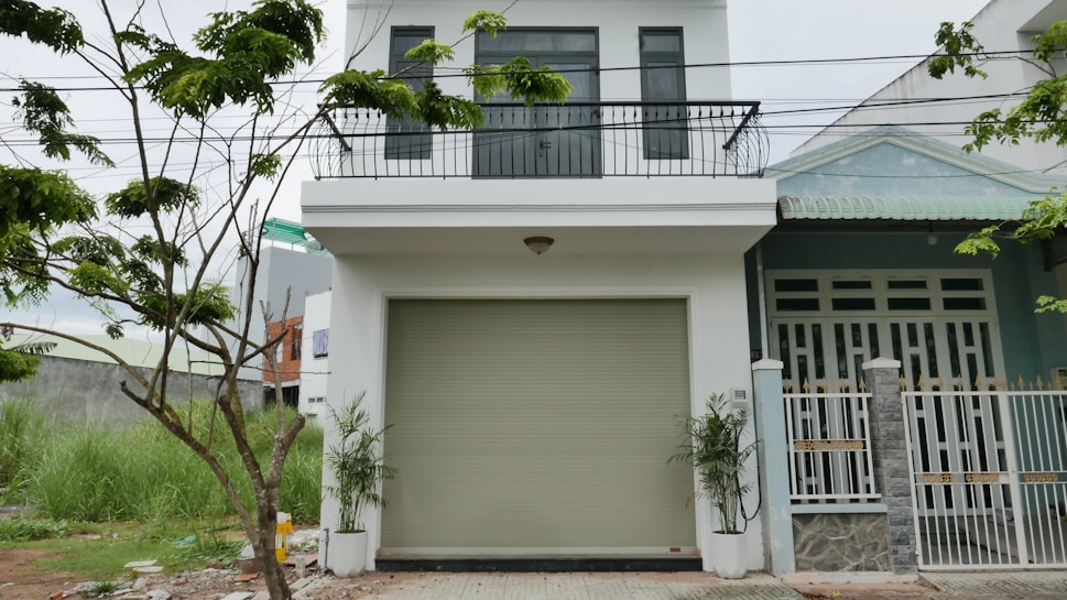 The image depicts a modern two-story house with a white facade and a balcony on the upper level. There is a large garage door in the center of the lower level. A small tree with green leaves is visible on the left side, and some grass grows near it. The house has a clean and minimalistic design.