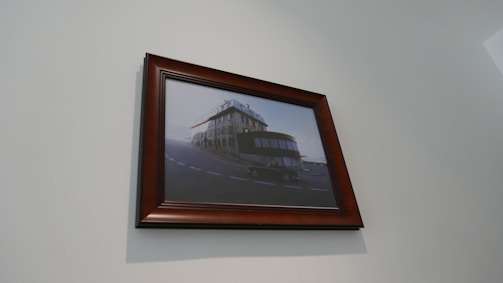 A framed painting of a surreal building that blends elements of both a traditional European-style house and a portion of a train. The artwork is hung on a light-colored wall.