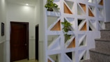 A modern interior space featuring a decorative partition wall with geometric cutouts, some of which are filled with small potted plants. The wall divides the space near a staircase with granite steps, and adjacent to a hallway with closed wooden doors. Soft lighting from above creates a warm and clean atmosphere.