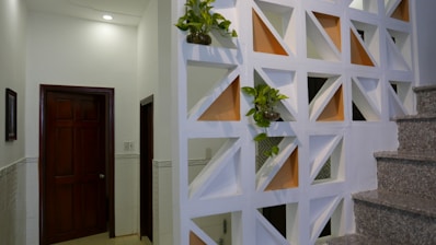 A modern interior space featuring a decorative partition wall with geometric cutouts, some of which are filled with small potted plants. The wall divides the space near a staircase with granite steps, and adjacent to a hallway with closed wooden doors. Soft lighting from above creates a warm and clean atmosphere.