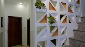 A modern interior space featuring a decorative partition wall with geometric cutouts, some of which are filled with small potted plants. The wall divides the space near a staircase with granite steps, and adjacent to a hallway with closed wooden doors. Soft lighting from above creates a warm and clean atmosphere.