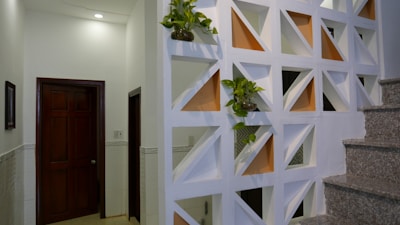 A modern interior space featuring a decorative partition wall with geometric cutouts, some of which are filled with small potted plants. The wall divides the space near a staircase with granite steps, and adjacent to a hallway with closed wooden doors. Soft lighting from above creates a warm and clean atmosphere.