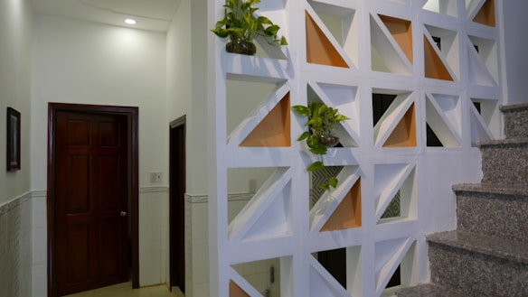 A modern interior space featuring a decorative partition wall with geometric cutouts, some of which are filled with small potted plants. The wall divides the space near a staircase with granite steps, and adjacent to a hallway with closed wooden doors. Soft lighting from above creates a warm and clean atmosphere.