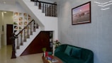 An interior view of a living area featuring a staircase with wooden railings leading upwards. The walls have a geometric design, and a framed picture hangs above a plush green velvet sofa. A small coffee table with red candles and a vase of flowers is positioned in front of the sofa. The space includes various decorative elements, plants, and a combination of wooden and tiled surfaces.