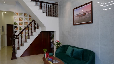 An interior view of a living area featuring a staircase with wooden railings leading upwards. The walls have a geometric design, and a framed picture hangs above a plush green velvet sofa. A small coffee table with red candles and a vase of flowers is positioned in front of the sofa. The space includes various decorative elements, plants, and a combination of wooden and tiled surfaces.