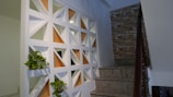 A stairway with gray stone steps and a wooden handrail is adjacent to a decorative wall featuring white geometric cutouts with orange backgrounds. Small plants are placed within some of the cutouts. The other side of the stairway has a wall with a textured, brick-like appearance.