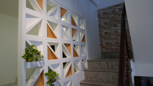 A stairway with gray stone steps and a wooden handrail is adjacent to a decorative wall featuring white geometric cutouts with orange backgrounds. Small plants are placed within some of the cutouts. The other side of the stairway has a wall with a textured, brick-like appearance.