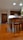 A well-lit kitchen area with wooden flooring and furniture. There are wooden cabinets on the wall, a dining table with chairs in the foreground, and a plant near the door. Two reflective pendant lights hang from the ceiling.