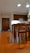 A well-lit kitchen area with wooden flooring and furniture. There are wooden cabinets on the wall, a dining table with chairs in the foreground, and a plant near the door. Two reflective pendant lights hang from the ceiling.