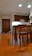 A well-lit kitchen area with wooden flooring and furniture. There are wooden cabinets on the wall, a dining table with chairs in the foreground, and a plant near the door. Two reflective pendant lights hang from the ceiling.