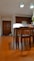 A well-lit kitchen area with wooden flooring and furniture. There are wooden cabinets on the wall, a dining table with chairs in the foreground, and a plant near the door. Two reflective pendant lights hang from the ceiling.