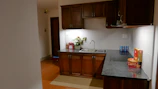 A small kitchen area featuring wooden cabinetry, a dark countertop, and modern appliances. A potted plant and a box of drinks are placed near the sink. On the countertop, there are various packaged food items, including tins and boxes.