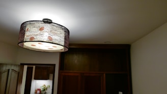 A modern room with a large decorative pendant light hanging from the ceiling, featuring a circular design with a patterned shade. The room has a dark wooden wardrobe and a partially visible kitchen area with white walls and cabinets. The lighting casts a warm, subtle glow.